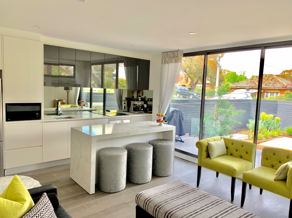 Small contemporary single-wall open plan kitchen in Melbourne with a built-in sink, shaker cabinets, white cabinets, marble worktops, grey splashback, mirror splashback, black appliances, light hardwood flooring, an island, grey floors and white worktops.
