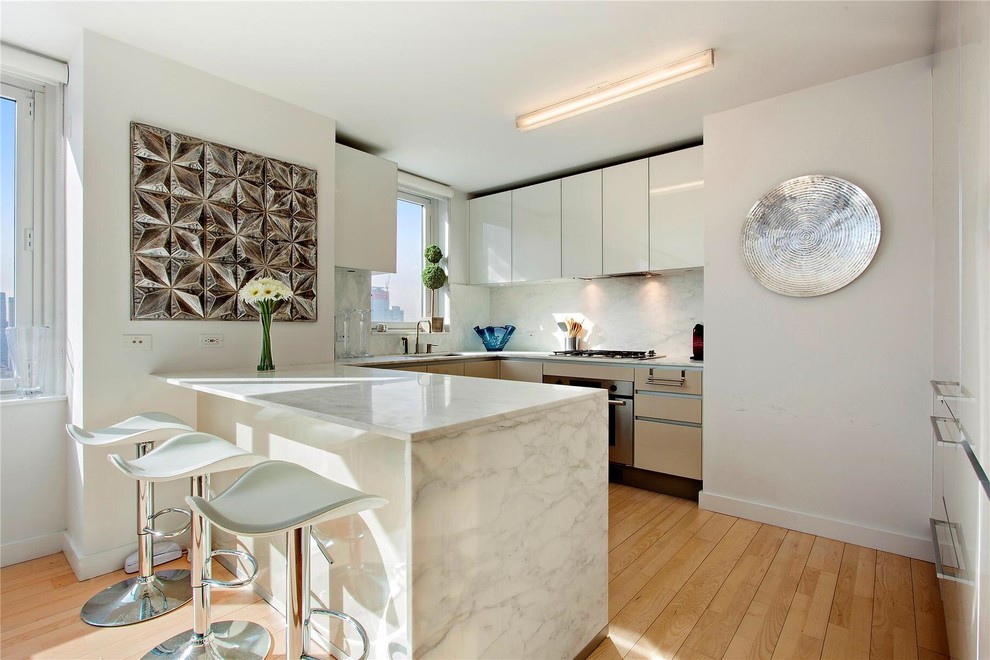 This is an example of a contemporary u-shaped kitchen in New York with flat-panel cabinets, white cabinets, white splashback, stone slab splashback, stainless steel appliances, medium hardwood flooring and a breakfast bar.