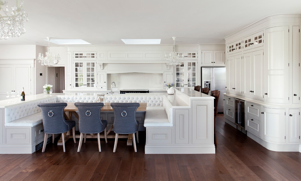 Klassisk inredning av ett stort kök, med luckor med glaspanel, bänkskiva i kvartsit, vitt stänkskydd, mörkt trägolv, vita skåp, rostfria vitvaror, en köksö och brunt golv