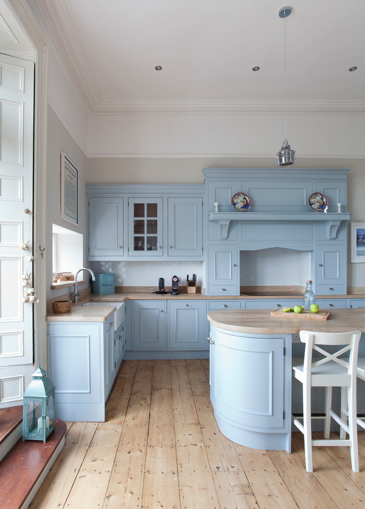 This is an example of a farmhouse l-shaped kitchen in Dublin with an island, a belfast sink, recessed-panel cabinets, blue cabinets, wood worktops, white splashback, metro tiled splashback, light hardwood flooring and beige floors.