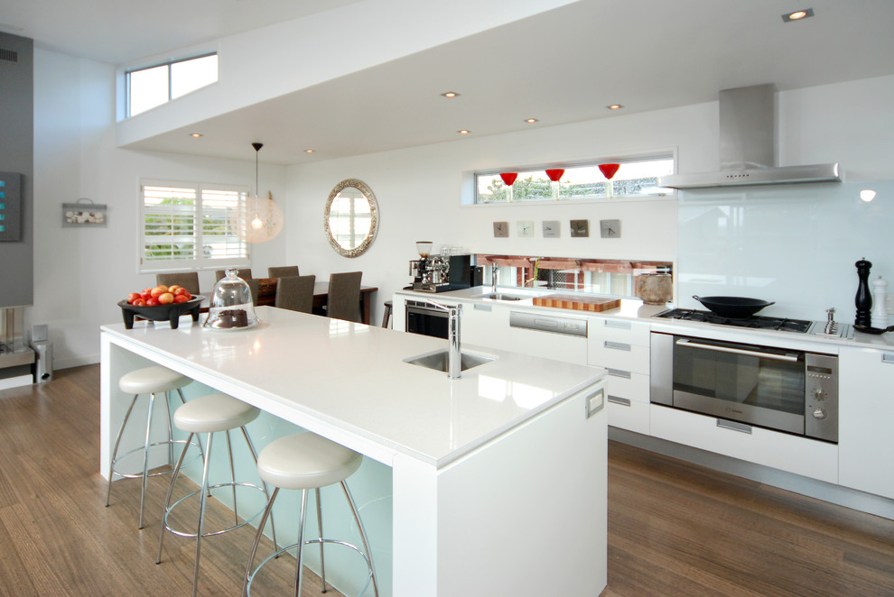 Einzeilige, Mittelgroße Moderne Wohnküche mit Waschbecken, flächenbündigen Schrankfronten, weißen Schränken, Granit-Arbeitsplatte, braunem Holzboden, Kücheninsel, Küchenrückwand in Weiß, Glasrückwand und Küchengeräten aus Edelstahl in Auckland