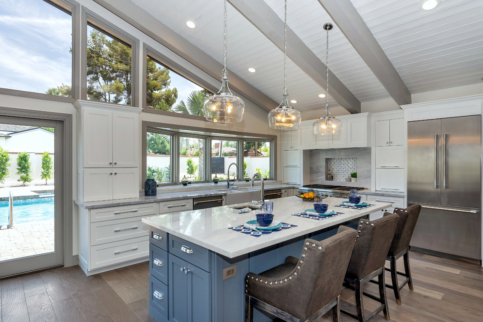 Example of a transitional l-shaped medium tone wood floor and brown floor kitchen design in Phoenix with a farmhouse sink, shaker cabinets, white cabinets, gray backsplash, stainless steel appliances, an island and white countertops