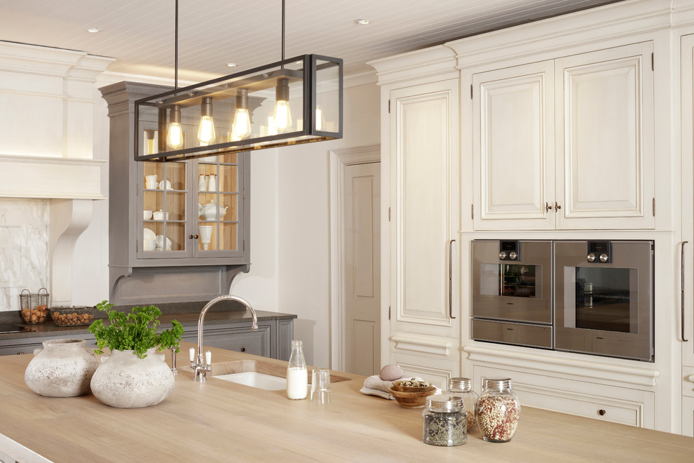 Inspiration for a large traditional kitchen/diner in Dublin with a double-bowl sink, raised-panel cabinets, distressed cabinets, wood worktops, white splashback, marble splashback, stainless steel appliances, limestone flooring, an island and beige floors.