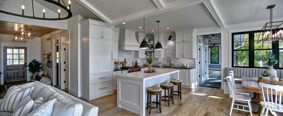 Foto på ett maritimt svart kök, med en undermonterad diskho, skåp i shakerstil, vita skåp, vitt stänkskydd, mellanmörkt trägolv, en köksö, brunt golv, bänkskiva i kvartsit, stänkskydd i marmor och integrerade vitvaror