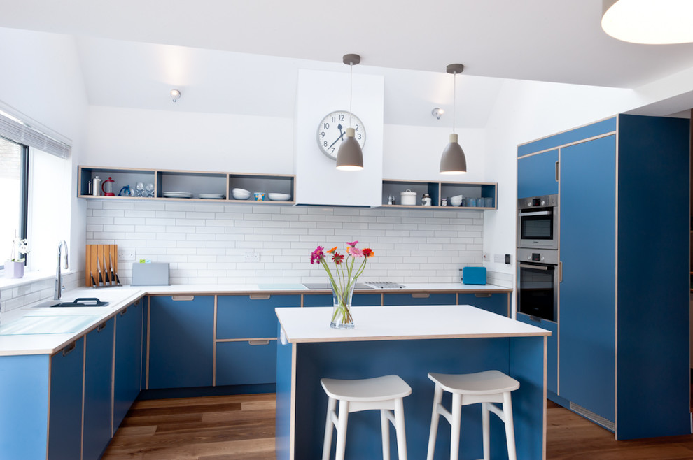Design ideas for a large contemporary u-shaped kitchen in Kent with flat-panel cabinets, blue cabinets, white splashback, metro tiled splashback, medium hardwood flooring and an island.
