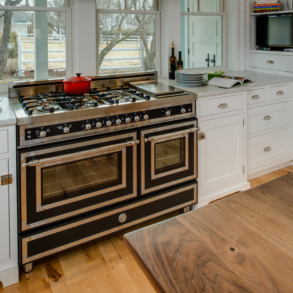 This is an example of a medium sized beach style u-shaped kitchen/diner in Other with a belfast sink, beaded cabinets, white cabinets, marble worktops, white splashback, window splashback, black appliances, light hardwood flooring, an island and beige floors.