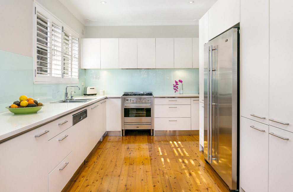 Modern inredning av ett avskilt, mellanstort l-kök, med en dubbel diskho, vita skåp, bänkskiva i kalksten, glaspanel som stänkskydd, rostfria vitvaror, släta luckor, blått stänkskydd och mellanmörkt trägolv