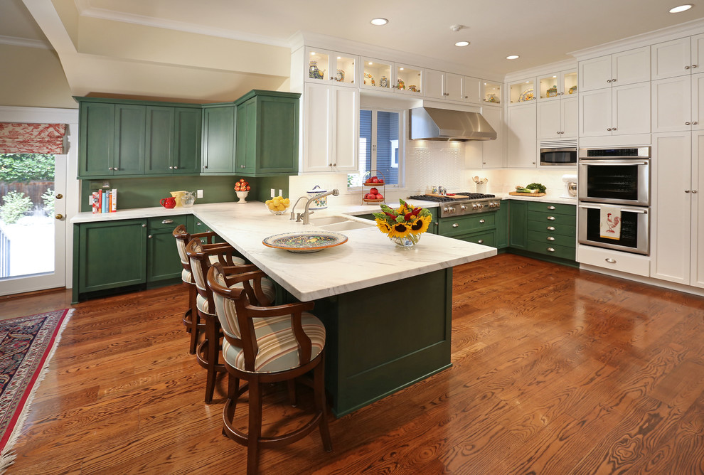Inredning av ett klassiskt stort kök, med en dubbel diskho, luckor med infälld panel, gröna skåp, bänkskiva i kvartsit, vitt stänkskydd, stänkskydd i keramik, rostfria vitvaror, mellanmörkt trägolv och en köksö