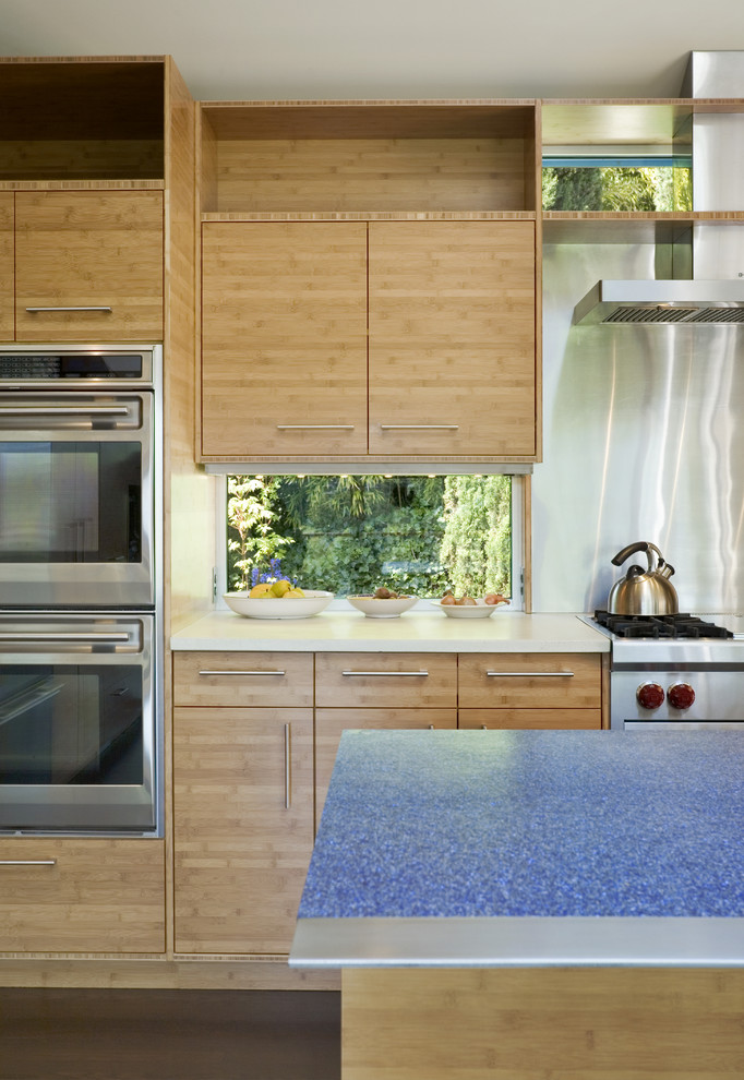 Exemple d'une cuisine ouverte tendance en bois clair et L avec un placard à porte plane, un plan de travail en surface solide, une crédence métallisée, un électroménager en acier inoxydable et îlot.