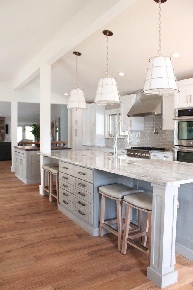 Example of a mid-sized transitional galley medium tone wood floor and brown floor kitchen design in St Louis with a farmhouse sink, recessed-panel cabinets, gray cabinets, gray backsplash, subway tile backsplash, stainless steel appliances and two islands