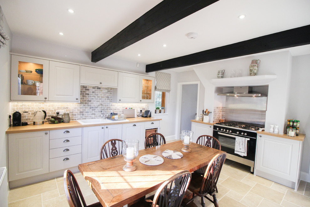 This is an example of a medium sized country l-shaped kitchen/diner in London with shaker cabinets, grey cabinets, wood worktops, black appliances, beige floors, an integrated sink, multi-coloured splashback, brick splashback and limestone flooring.