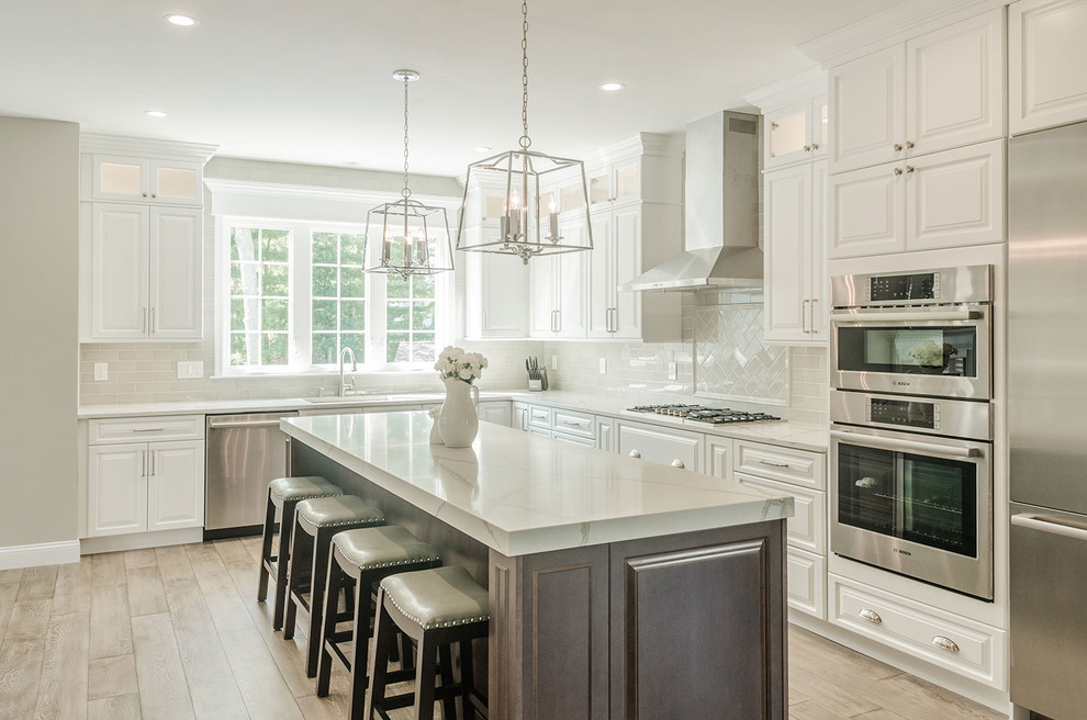 Example of a classic l-shaped light wood floor and beige floor kitchen design in Philadelphia with an island, raised-panel cabinets, white cabinets, white backsplash, subway tile backsplash, stainless steel appliances and white countertops