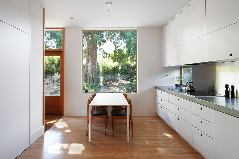 Inredning av ett modernt mellanstort kök, med mellanmörkt trägolv, släta luckor, vita skåp, bänkskiva i rostfritt stål, fönster som stänkskydd och brunt golv