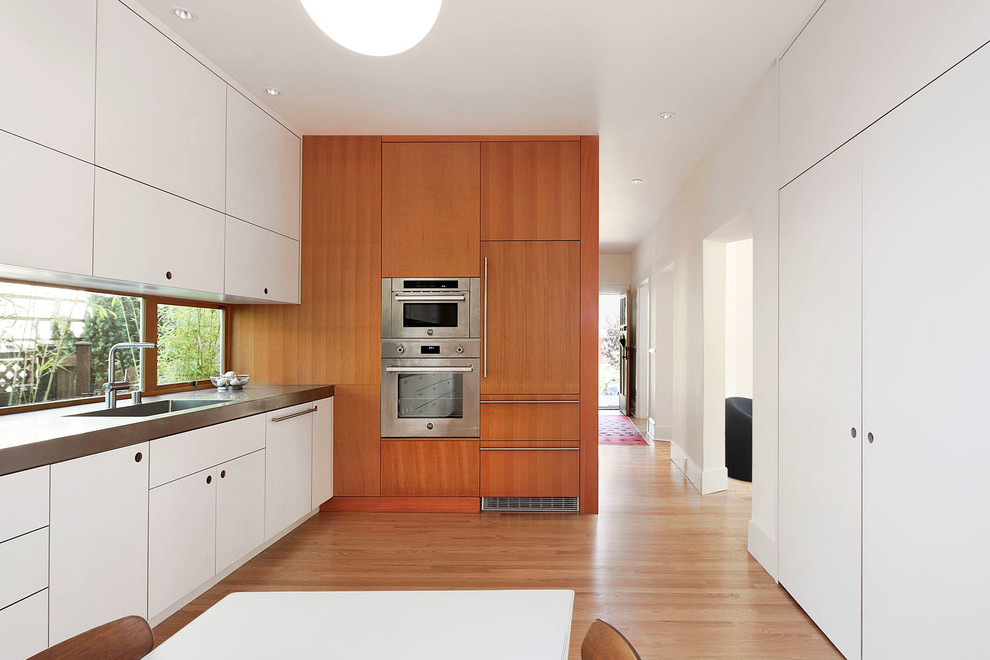 Modern inredning av ett mellanstort l-kök, med släta luckor, vita skåp, rostfria vitvaror, ljust trägolv, en undermonterad diskho, fönster som stänkskydd och brunt golv