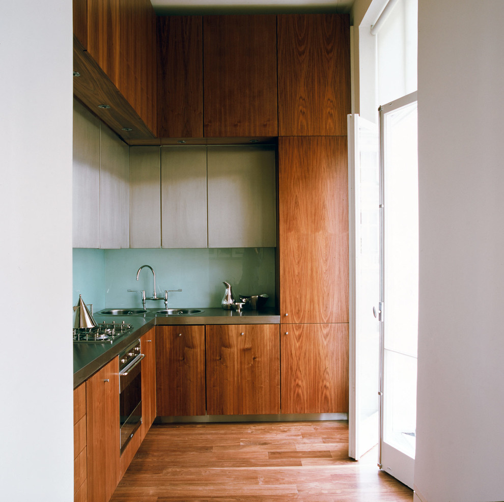 Exempel på ett avskilt, litet retro l-kök, med en nedsänkt diskho, släta luckor, skåp i mellenmörkt trä, bänkskiva i rostfritt stål, grönt stänkskydd, glaspanel som stänkskydd, rostfria vitvaror och mellanmörkt trägolv