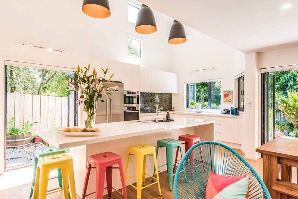 Zweizeilige, Große Moderne Wohnküche mit weißen Schränken, Kücheninsel, flächenbündigen Schrankfronten, Küchenrückwand in Schwarz, Küchengeräten aus Edelstahl, braunem Holzboden und beigem Boden in Auckland