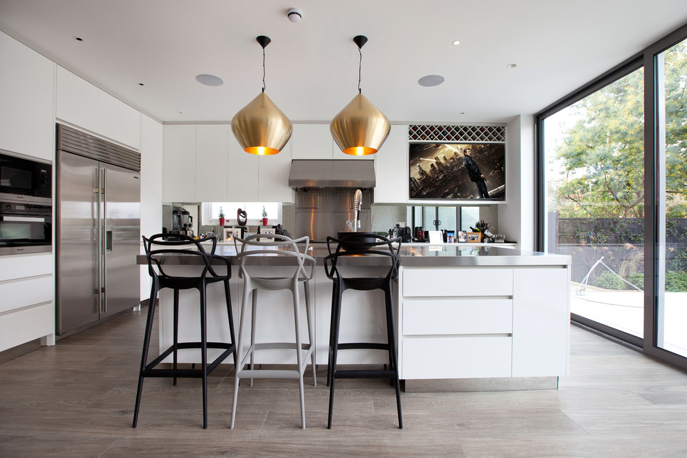 This is an example of a large contemporary l-shaped open plan kitchen in London with flat-panel cabinets, white cabinets, stainless steel worktops, stainless steel appliances, light hardwood flooring and an island.