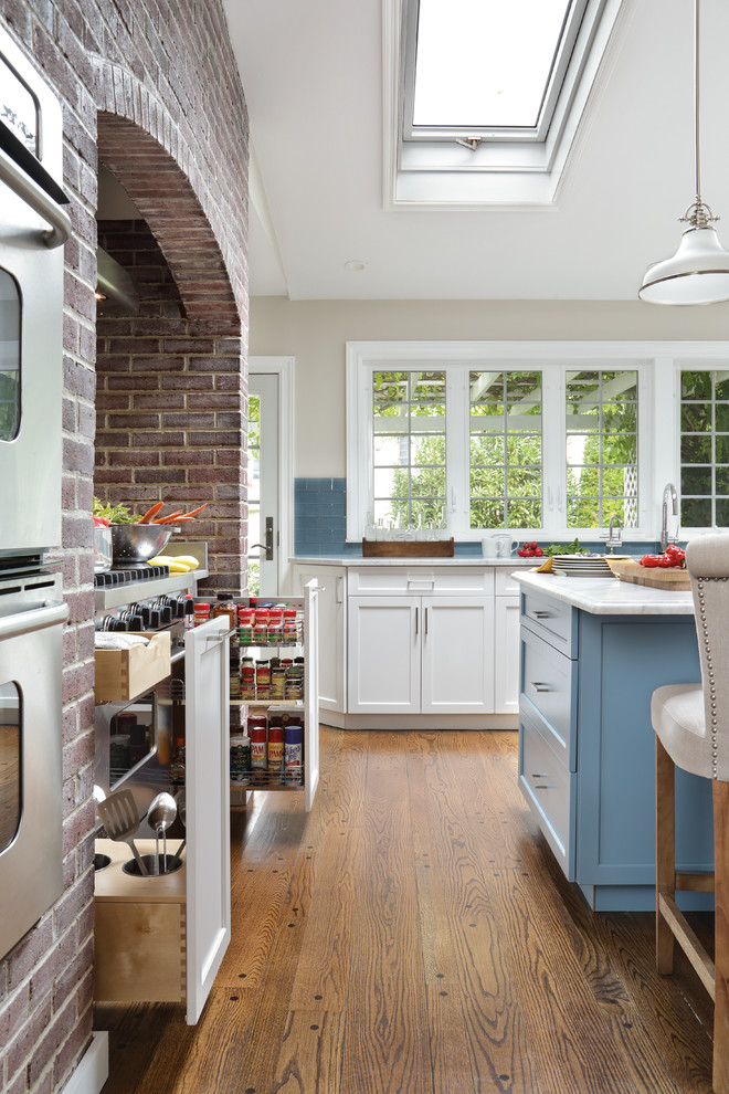 Inredning av ett klassiskt stort kök, med en undermonterad diskho, luckor med infälld panel, vita skåp, blått stänkskydd, stänkskydd i glaskakel, rostfria vitvaror, mellanmörkt trägolv och en köksö
