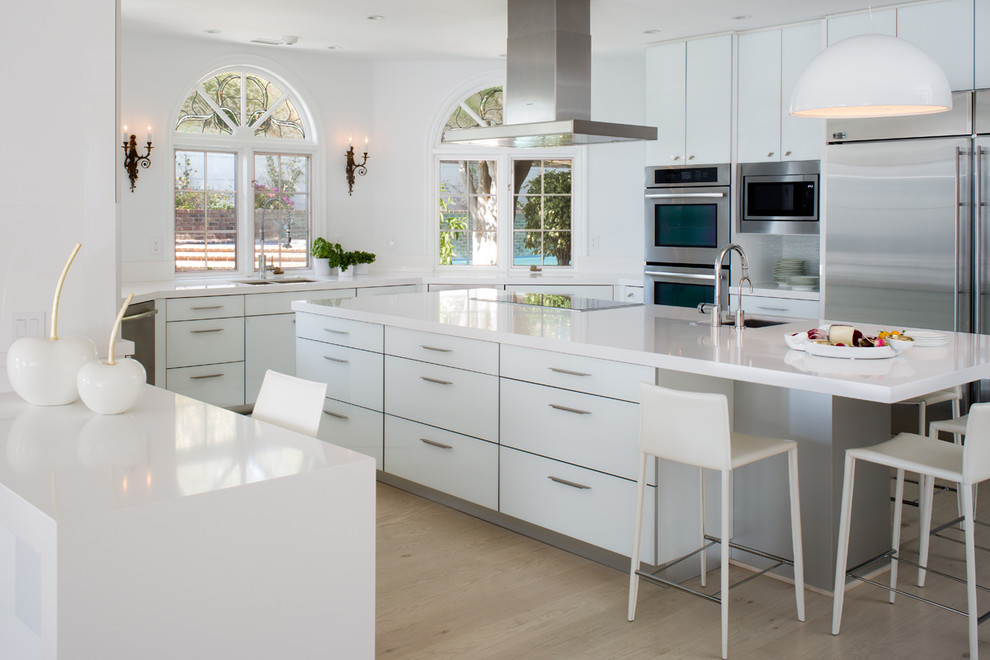 This is an example of a contemporary u-shaped kitchen/diner in San Diego with a submerged sink, flat-panel cabinets, white cabinets, stainless steel appliances, an island, composite countertops, white splashback and light hardwood flooring.