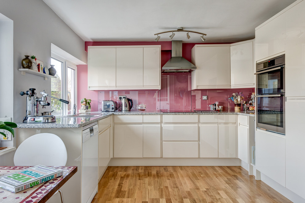 Design ideas for a small u-shaped enclosed kitchen in Devon with a submerged sink, flat-panel cabinets, white cabinets, granite worktops, pink splashback, glass sheet splashback, stainless steel appliances and light hardwood flooring.