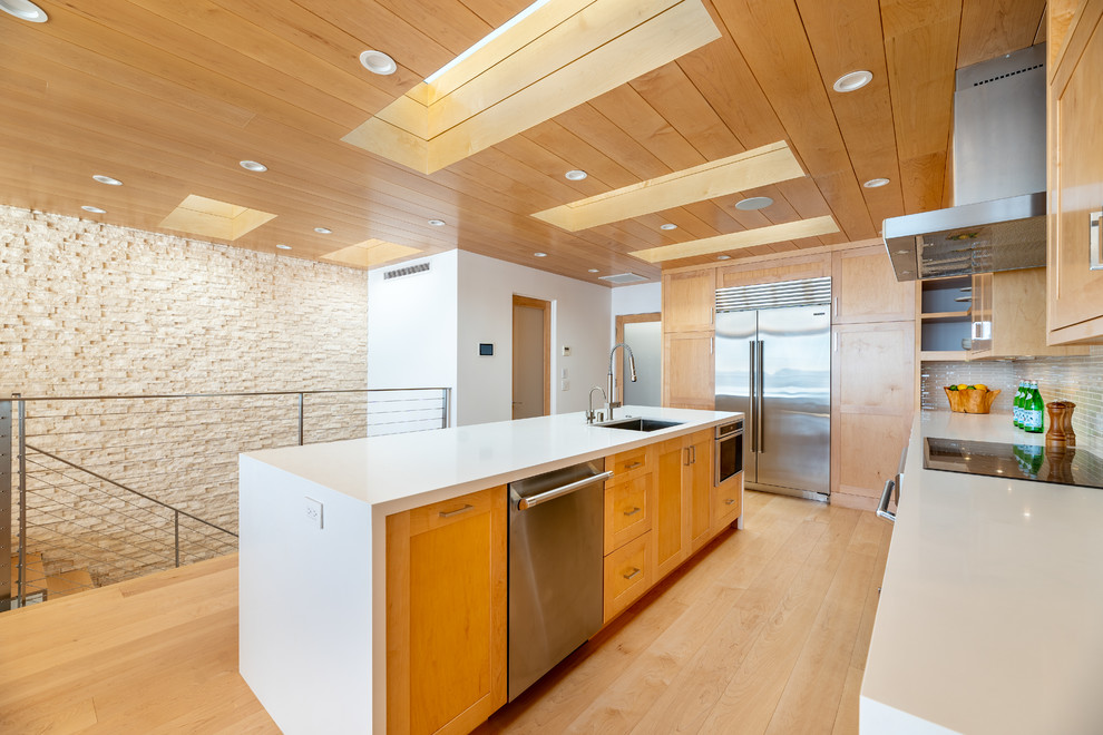 Inspiration for an expansive contemporary galley open plan kitchen in Los Angeles with a single-bowl sink, shaker cabinets, light wood cabinets, engineered stone countertops, beige splashback, glass tiled splashback, stainless steel appliances, light hardwood flooring, an island, yellow floors and white worktops.
