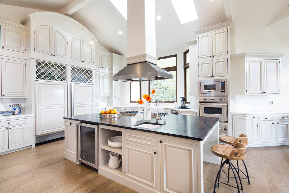 Example of a large classic u-shaped light wood floor eat-in kitchen design in San Francisco with raised-panel cabinets, white cabinets, white backsplash, stainless steel appliances, an island, a farmhouse sink, soapstone countertops and stone slab backsplash