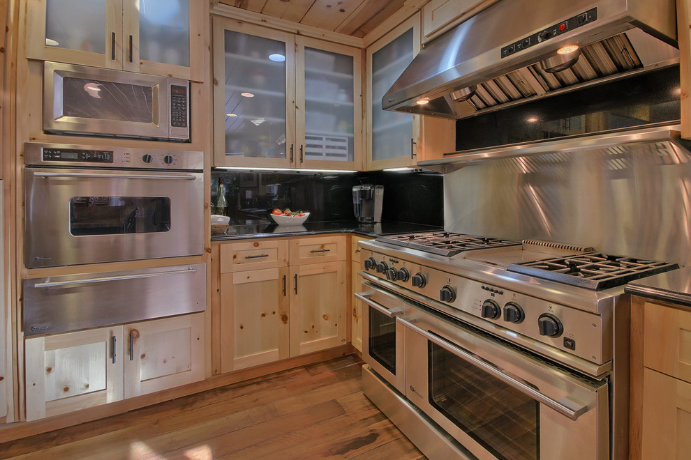 他の地域にある広いラスティックスタイルのおしゃれなキッチン (アンダーカウンターシンク、ガラス扉のキャビネット、淡色木目調キャビネット、シルバーの調理設備、無垢フローリング、黒いキッチンパネル) の写真