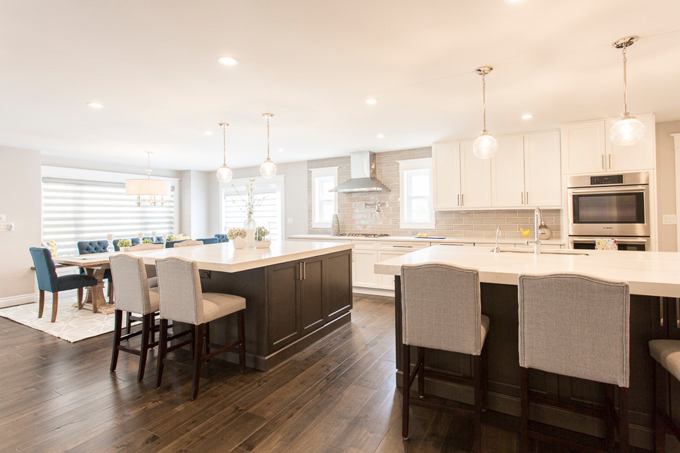 Photo of a large classic l-shaped kitchen/diner in Denver with a single-bowl sink, recessed-panel cabinets, white cabinets, engineered stone countertops, grey splashback, ceramic splashback, stainless steel appliances, medium hardwood flooring, multiple islands and brown floors.