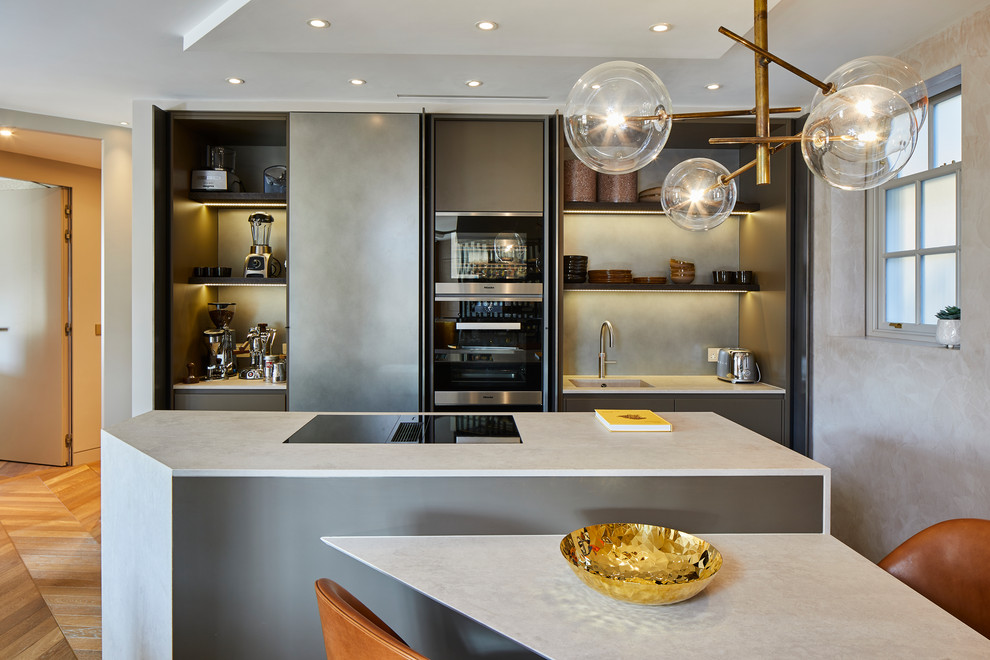 Moderne Wohnküche mit integriertem Waschbecken, grauen Schränken, Küchenrückwand in Grau, schwarzen Elektrogeräten, braunem Holzboden, zwei Kücheninseln und grauer Arbeitsplatte in Kent