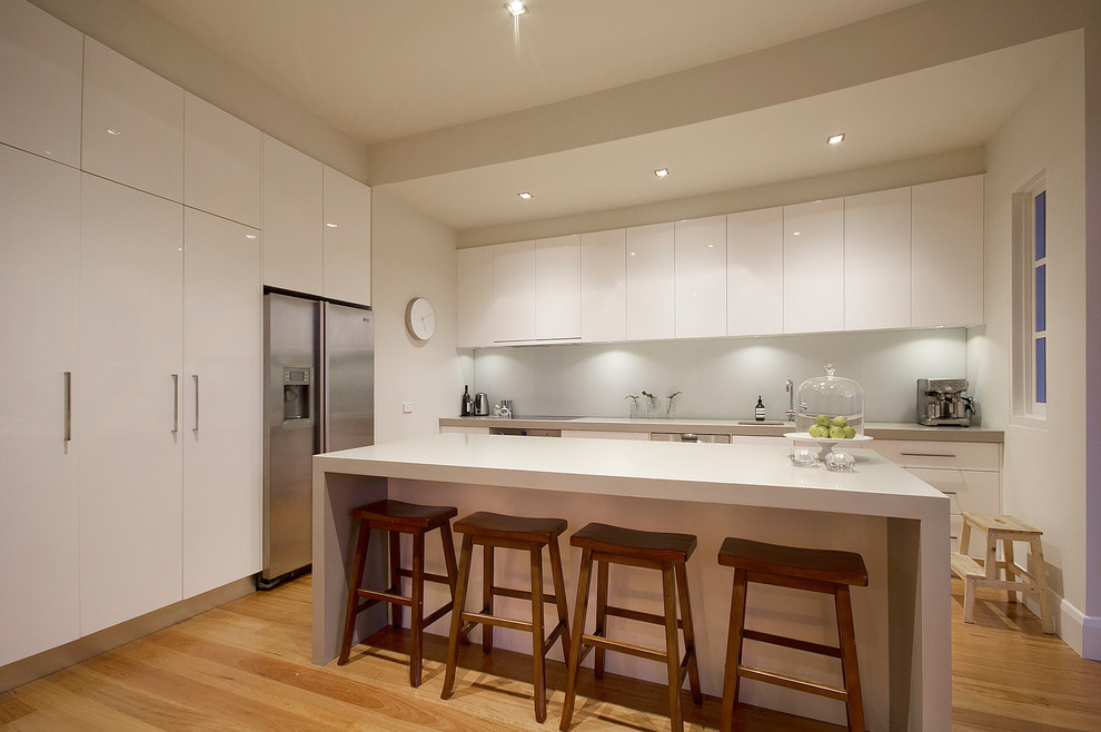 Mittelgroße Moderne Wohnküche in L-Form mit Unterbauwaschbecken, flächenbündigen Schrankfronten, weißen Schränken, Küchenrückwand in Weiß, Küchengeräten aus Edelstahl, hellem Holzboden und Kücheninsel in Melbourne