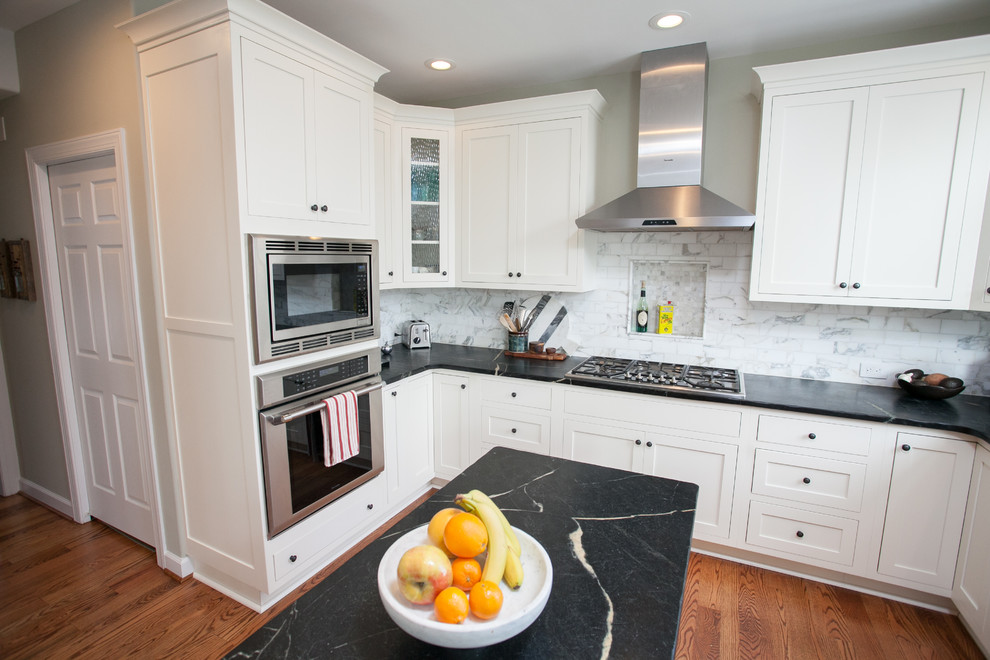 Modelo de cocina clásica renovada, de tamaño medio, con fregadero sobremueble, armarios estilo shaker, puertas de armario blancas, encimera de esteatita, salpicadero blanco, salpicadero de azulejos de piedra, electrodomésticos de acero inoxidable, suelo de madera en tonos medios y una isla