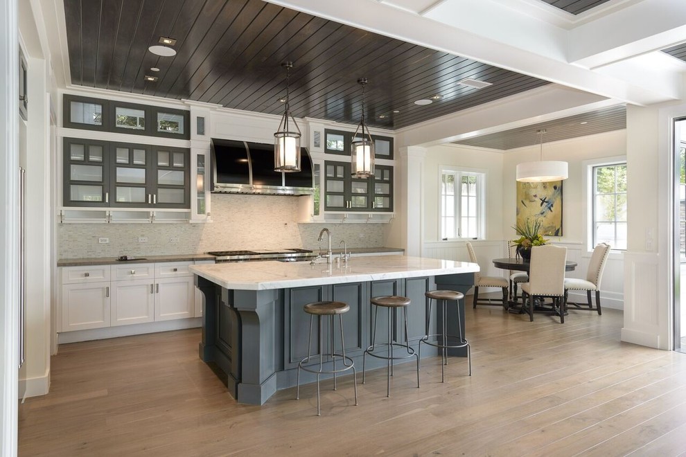 This is an example of a coastal kitchen/diner in Los Angeles with glass-front cabinets, grey cabinets, marble worktops, white splashback, light hardwood flooring, an island and beige floors.