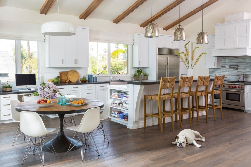 This is an example of a beach style l-shaped kitchen/diner in Los Angeles with shaker cabinets, white cabinets, an island, a belfast sink, multi-coloured splashback, matchstick tiled splashback, stainless steel appliances, dark hardwood flooring, brown floors and black worktops.
