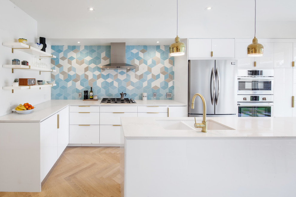 Bild på ett funkis l-kök, med en undermonterad diskho, släta luckor, vita skåp, bänkskiva i kvarts, blått stänkskydd, rostfria vitvaror, ljust trägolv och en köksö
