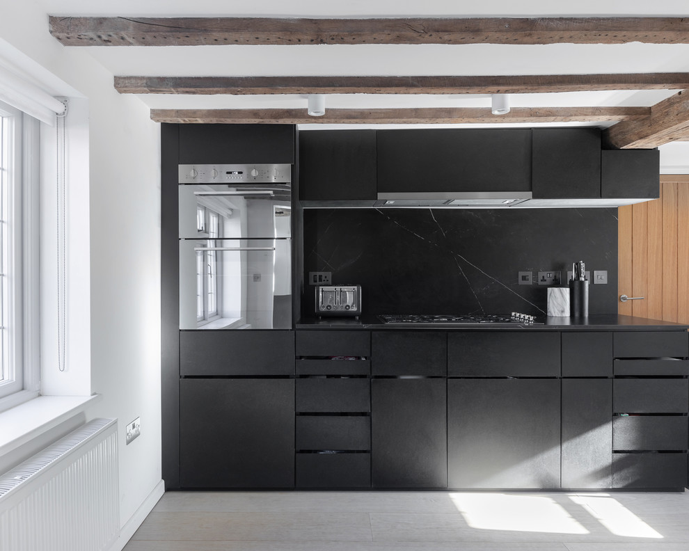 Example of a mid-sized cottage l-shaped light wood floor and beige floor open concept kitchen design in Oxfordshire with a drop-in sink, flat-panel cabinets, black cabinets, marble countertops, black backsplash, marble backsplash, stainless steel appliances, no island and black countertops