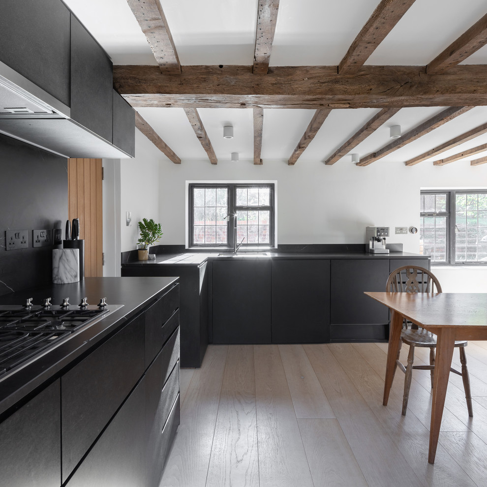 This is an example of a medium sized country l-shaped kitchen/diner in Oxfordshire with flat-panel cabinets, black cabinets, black splashback, light hardwood flooring, beige floors, black worktops and a submerged sink.