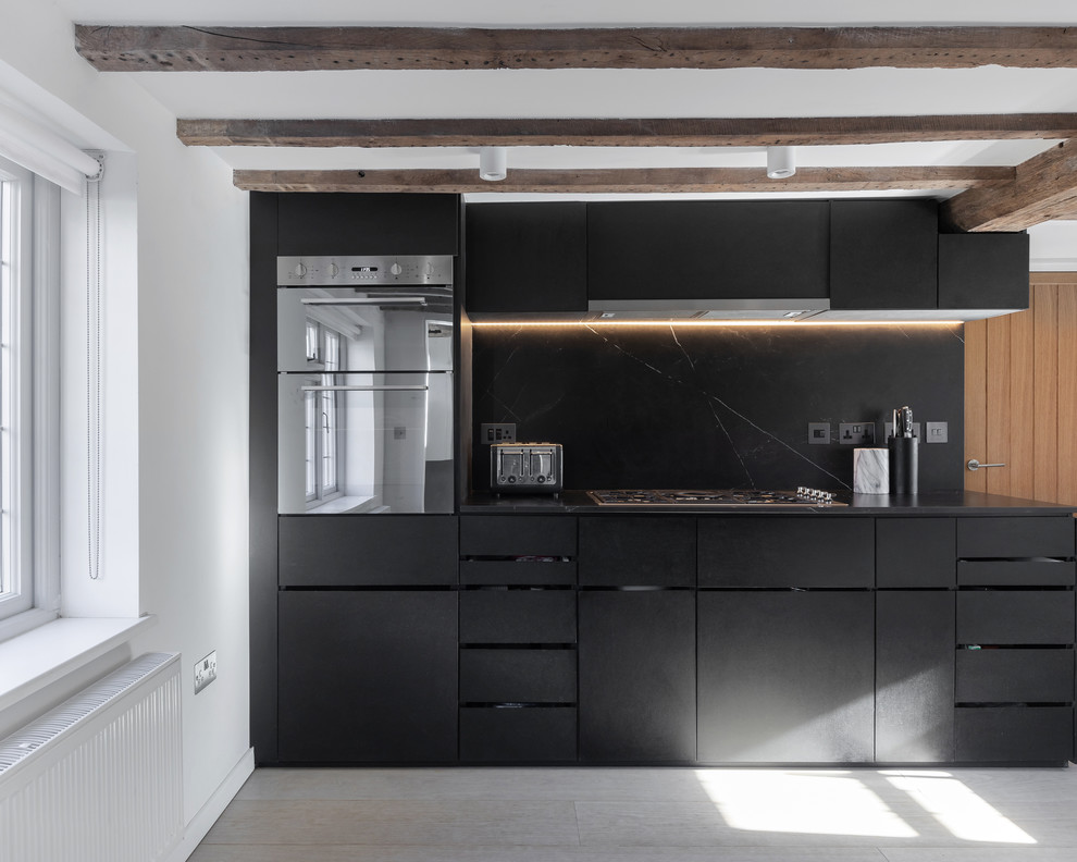 This is an example of a medium sized farmhouse l-shaped open plan kitchen in Oxfordshire with a built-in sink, flat-panel cabinets, black cabinets, marble worktops, black splashback, marble splashback, stainless steel appliances, light hardwood flooring, no island, beige floors and black worktops.