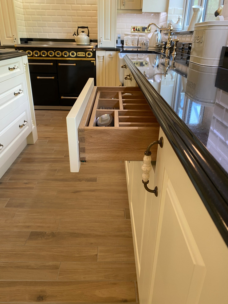 This is an example of a large classic cream and black u-shaped kitchen/diner in Sussex with a belfast sink, raised-panel cabinets, white cabinets, granite worktops, black splashback, granite splashback, integrated appliances, ceramic flooring, an island, multi-coloured floors, black worktops and feature lighting.