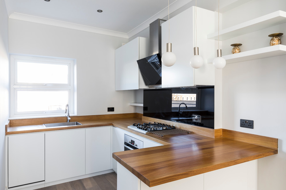 This is an example of a small contemporary u-shaped open plan kitchen in London with a built-in sink, flat-panel cabinets, white cabinets, wood worktops, black splashback, glass sheet splashback, stainless steel appliances, light hardwood flooring and no island.