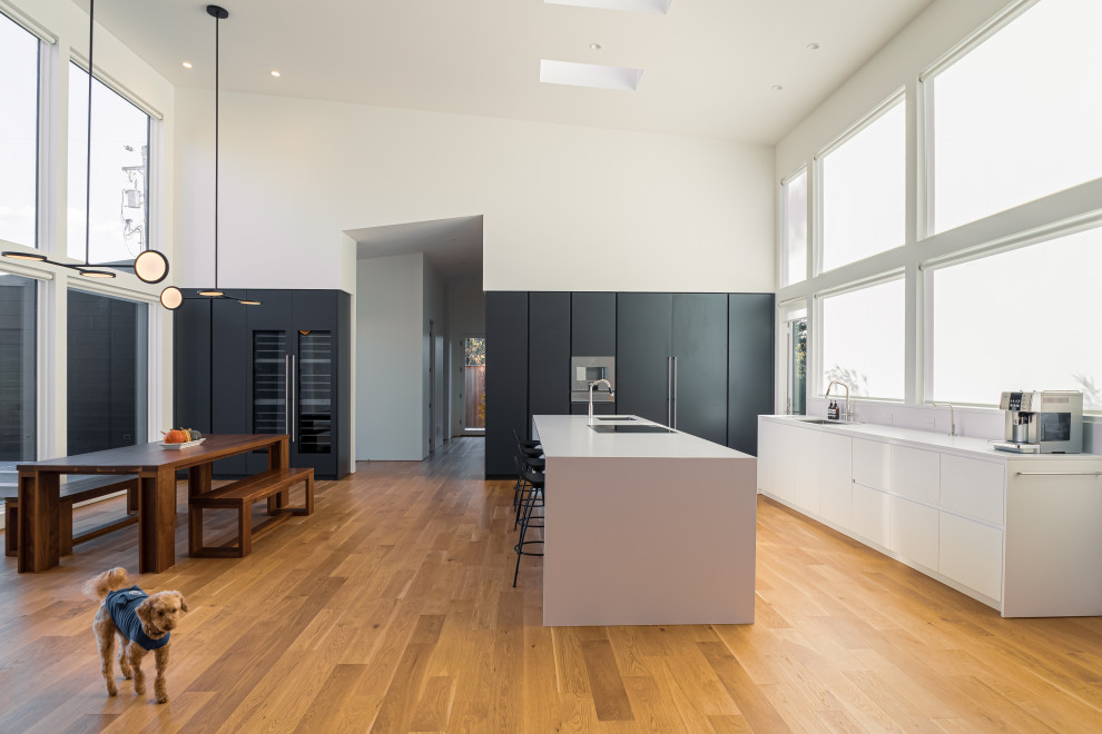 Photo of an expansive contemporary l-shaped open plan kitchen in Portland with a submerged sink, flat-panel cabinets, white cabinets, engineered stone countertops, white splashback, engineered quartz splashback, integrated appliances, light hardwood flooring, an island, brown floors and white worktops.