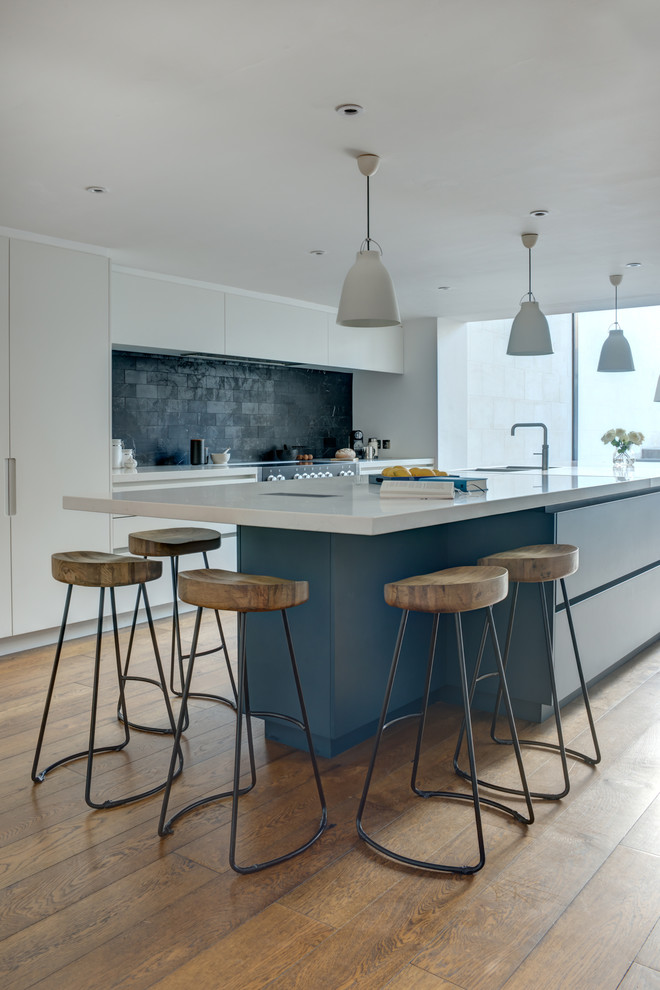 This is an example of an expansive contemporary single-wall open plan kitchen in London with an integrated sink, flat-panel cabinets, white cabinets, quartz worktops, black splashback, marble splashback, integrated appliances, medium hardwood flooring, an island, beige floors and white worktops.
