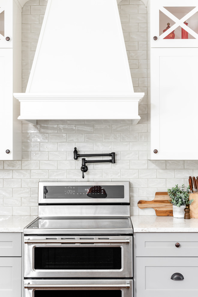 Imagen de cocina comedor de estilo de casa de campo grande con fregadero bajoencimera, armarios estilo shaker, puertas de armario blancas, encimera de cuarzo compacto, salpicadero blanco, salpicadero de azulejos de porcelana, electrodomésticos de acero inoxidable, suelo de madera en tonos medios, una isla, suelo marrón y encimeras blancas