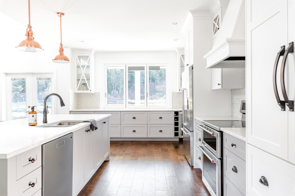 Example of a large farmhouse medium tone wood floor and brown floor eat-in kitchen design in Other with an undermount sink, shaker cabinets, white cabinets, quartz countertops, white backsplash, porcelain backsplash, stainless steel appliances, an island and white countertops