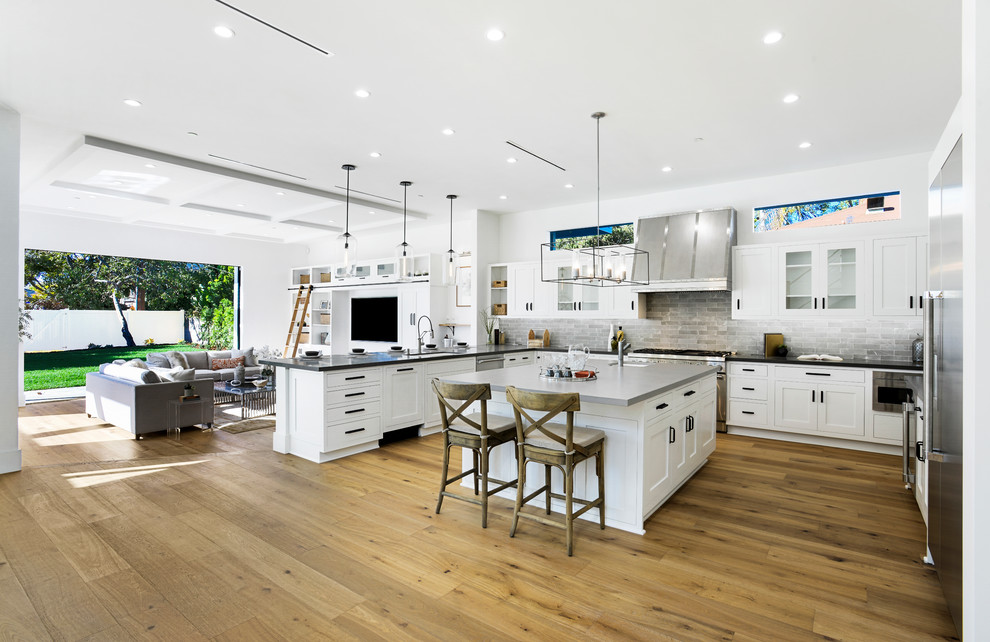 Inspiration for a large farmhouse u-shaped open plan kitchen in Los Angeles with a submerged sink, shaker cabinets, white cabinets, engineered stone countertops, grey splashback, porcelain splashback, stainless steel appliances, medium hardwood flooring, an island, grey worktops and brown floors.