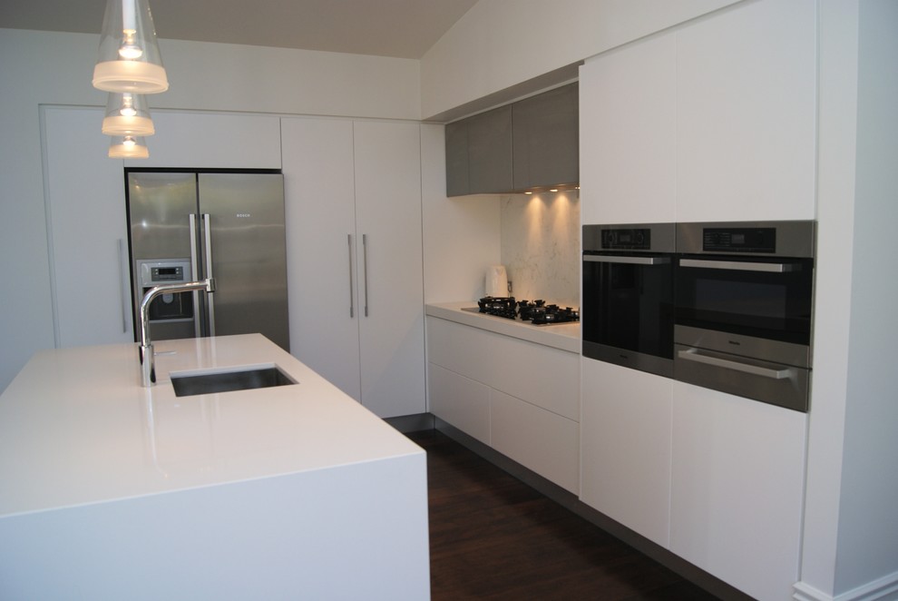 Photo of a small contemporary l-shaped kitchen/diner in Auckland with a submerged sink, flat-panel cabinets, engineered stone countertops, stone slab splashback, stainless steel appliances, dark hardwood flooring, an island and white cabinets.
