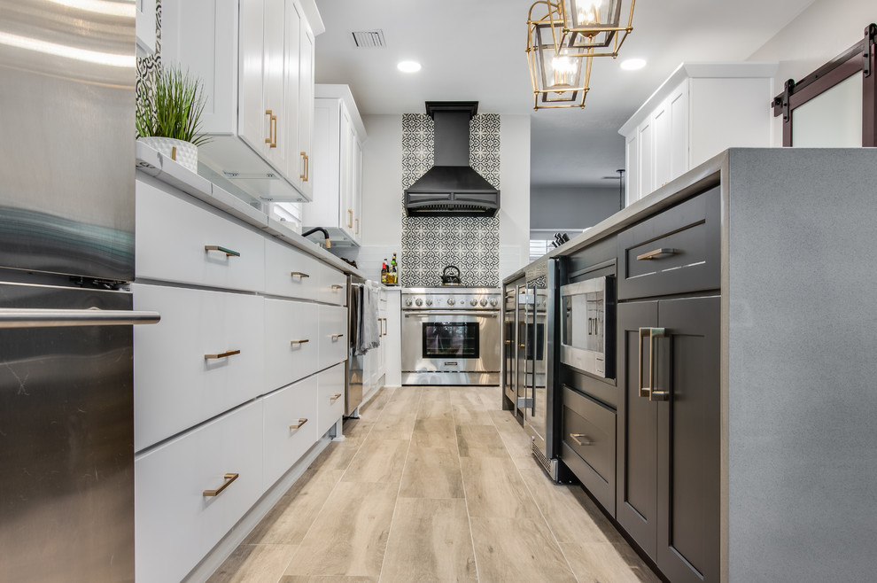Large traditional l-shaped kitchen pantry in Houston with a belfast sink, shaker cabinets, white cabinets, quartz worktops, white splashback, glass tiled splashback, stainless steel appliances, porcelain flooring, an island, grey floors and white worktops.