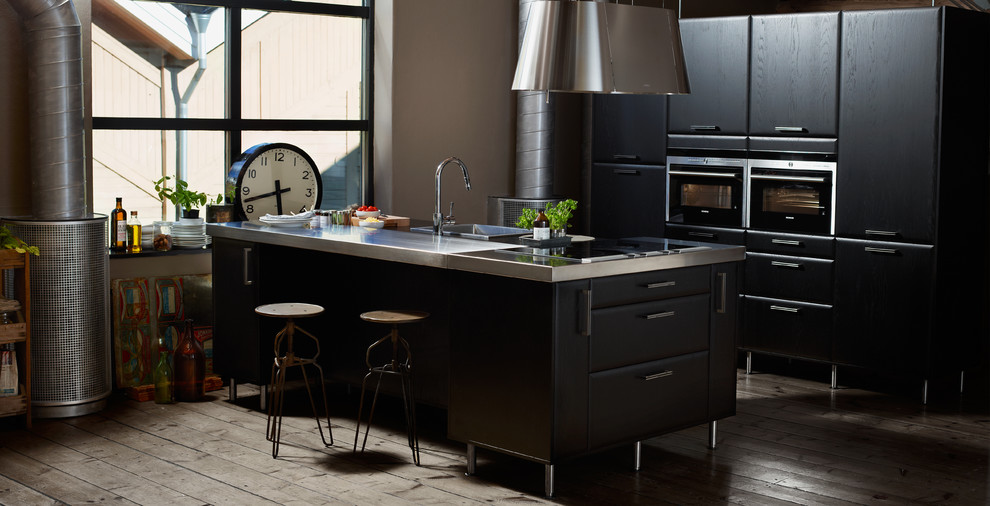 Diseño de cocina comedor moderna grande con fregadero encastrado, armarios con paneles lisos, puertas de armario negras, electrodomésticos de acero inoxidable, suelo de madera en tonos medios y una isla
