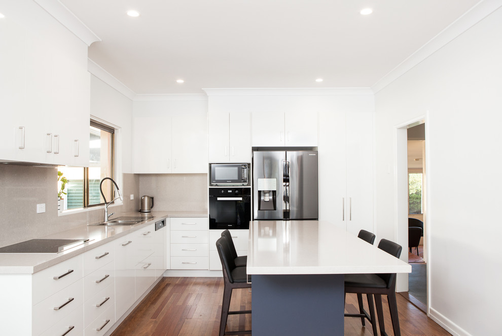 This is an example of a modern l-shaped kitchen pantry in Brisbane with a built-in sink, flat-panel cabinets, white cabinets, engineered stone countertops, grey splashback, stone slab splashback, stainless steel appliances, medium hardwood flooring, an island and brown floors.