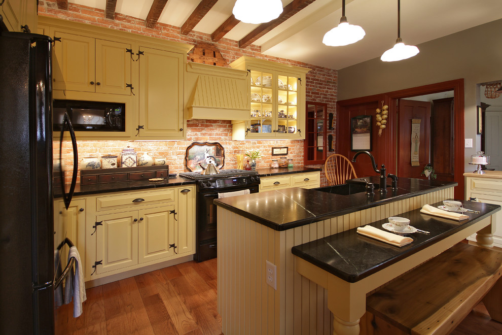 Inspiration for a small country l-shaped light wood floor enclosed kitchen remodel in Philadelphia with a farmhouse sink, beaded inset cabinets, yellow cabinets, soapstone countertops, red backsplash, black appliances and an island