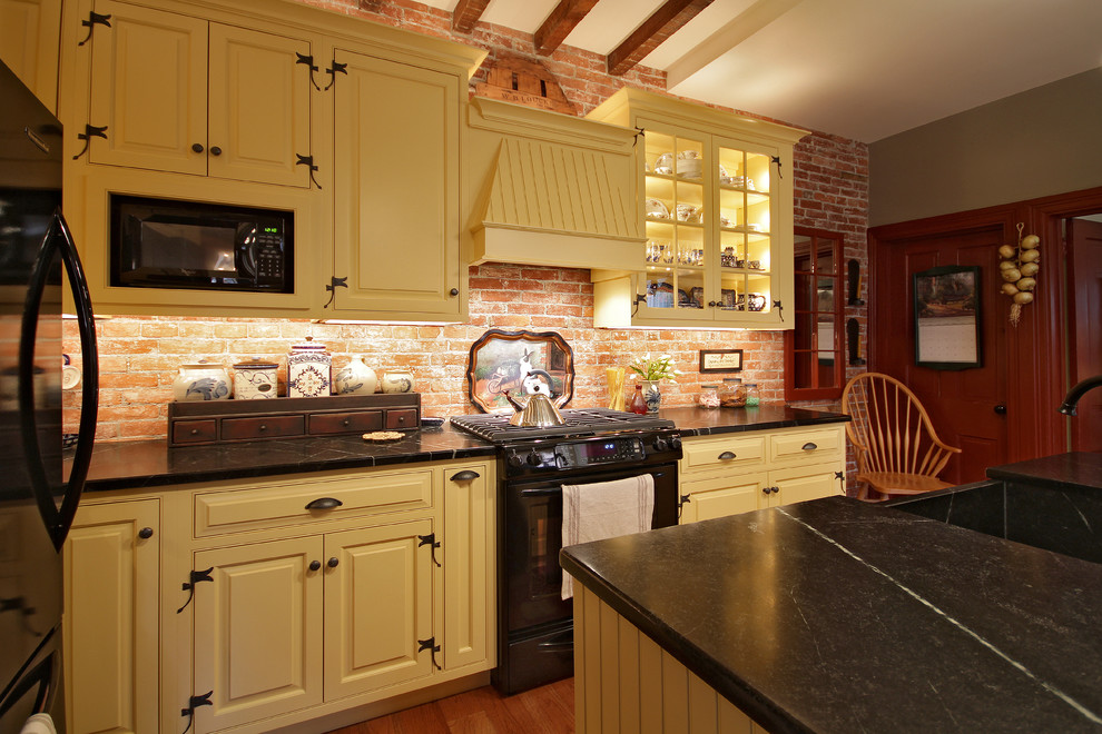 Bild på ett avskilt, litet lantligt l-kök, med en rustik diskho, luckor med profilerade fronter, gula skåp, bänkskiva i täljsten, rött stänkskydd, svarta vitvaror, ljust trägolv och en köksö